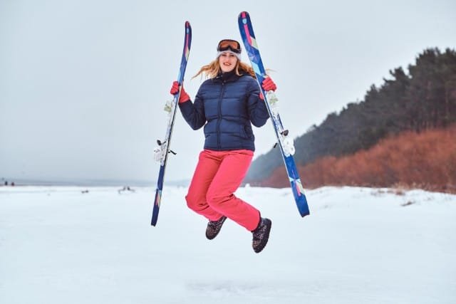 A woman in a red jacket and blue pants jumps joyfully in the air while skiing, showcasing her enthusiasm for the sport.