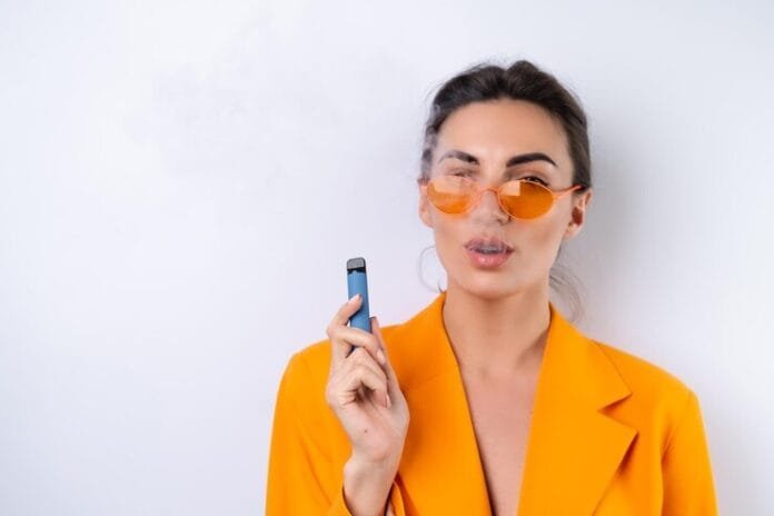 young-woman-trendy-stylish-glasses-bright-orange-oversized-jacket-white-background-smokes-electronic-cigarette_343596-8182 A woman dressed in an orange suit is holding a Fume vape, smiling as she poses for the photo.